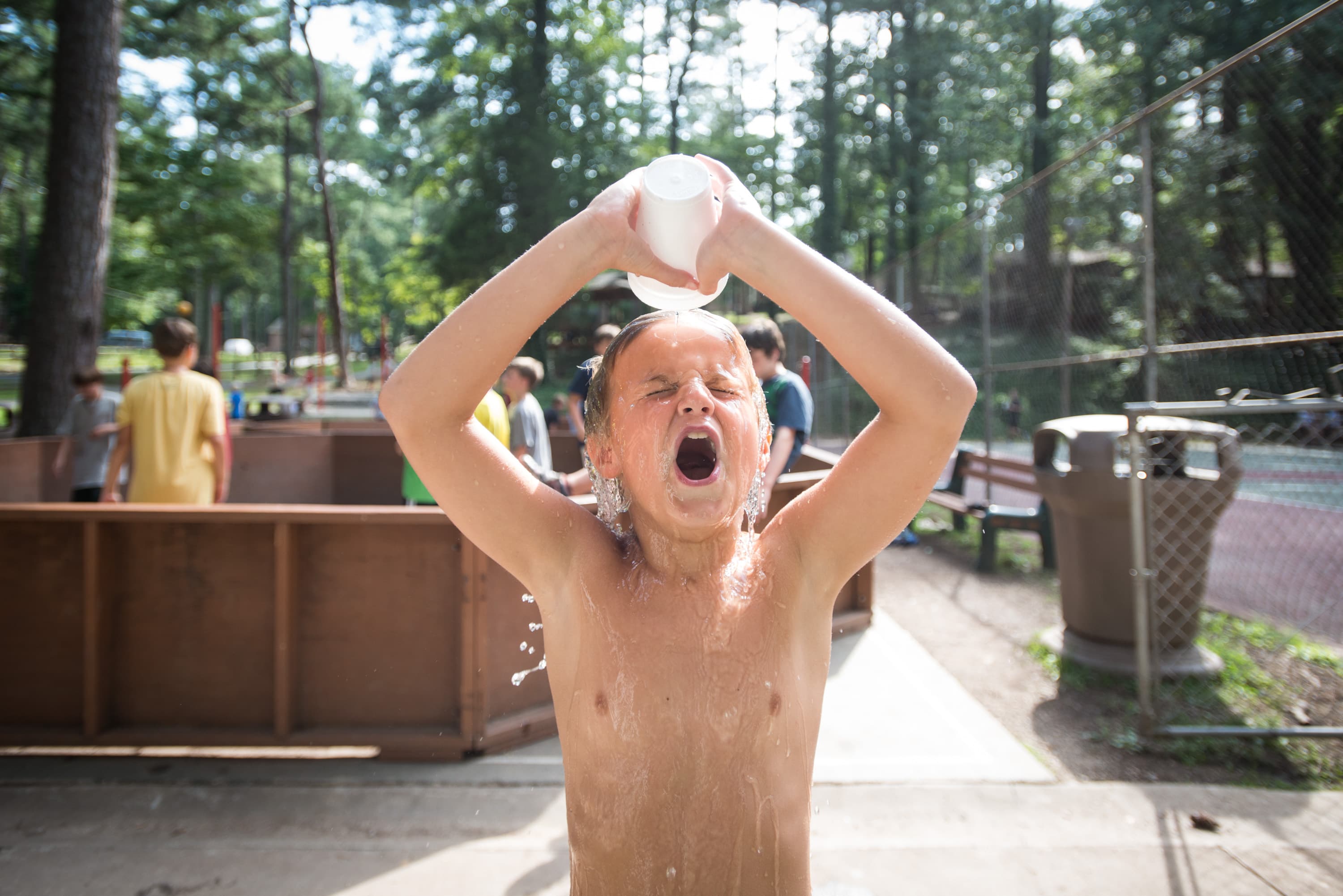 A child pouring water over their head from a cup, with their eyes closed and mouth open, standing in an outdoor area with other people and trees in the background.