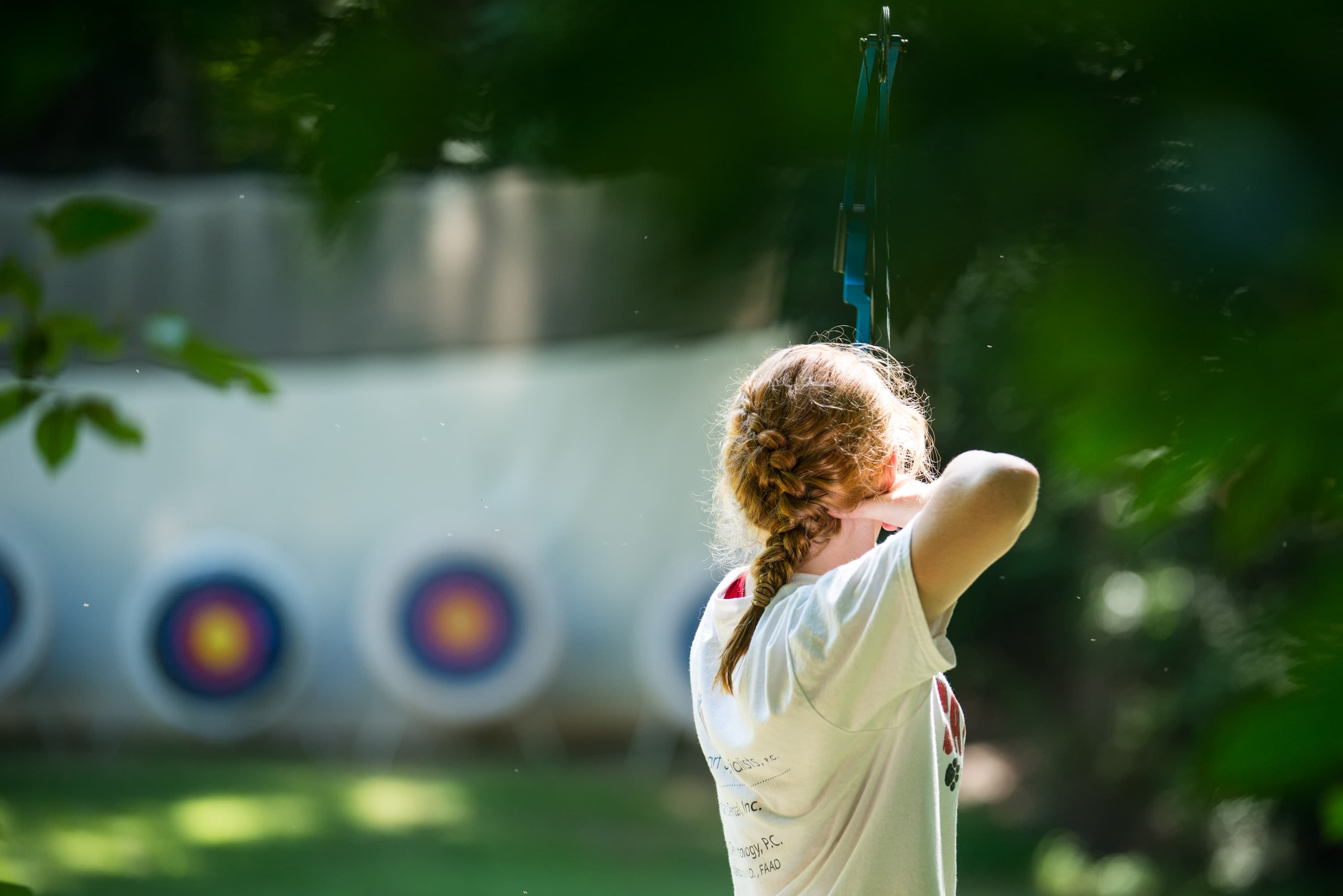 A person with braided hair, wearing a t-shirt, aiming a bow at archery targets in the background.