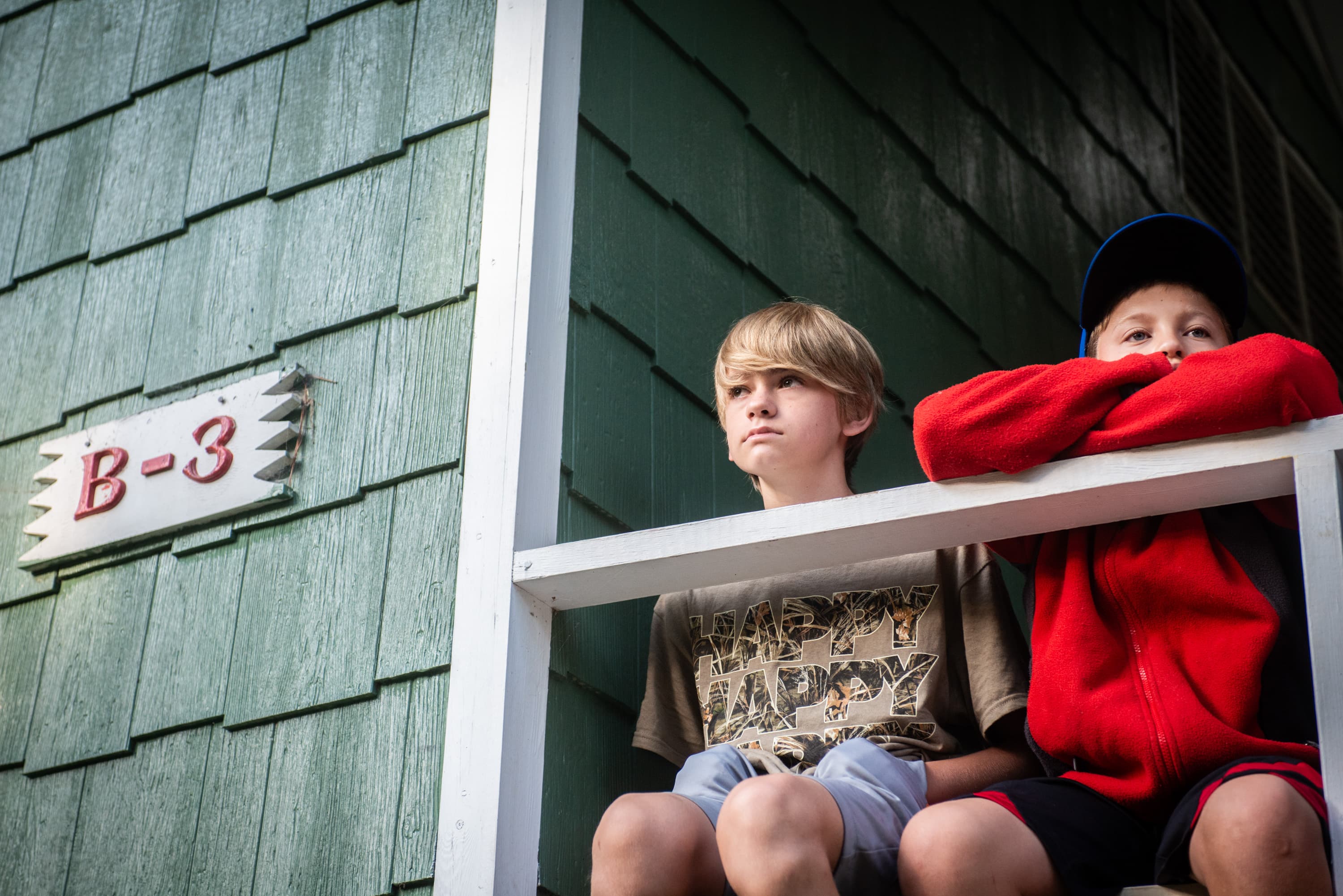 Two boys sitting on a porch. One is wearing a shirt with the word 'HAPPY' printed on it, and the other is wearing a red jacket and a blue cap. Above them on the wall is a sign that reads "B-3."