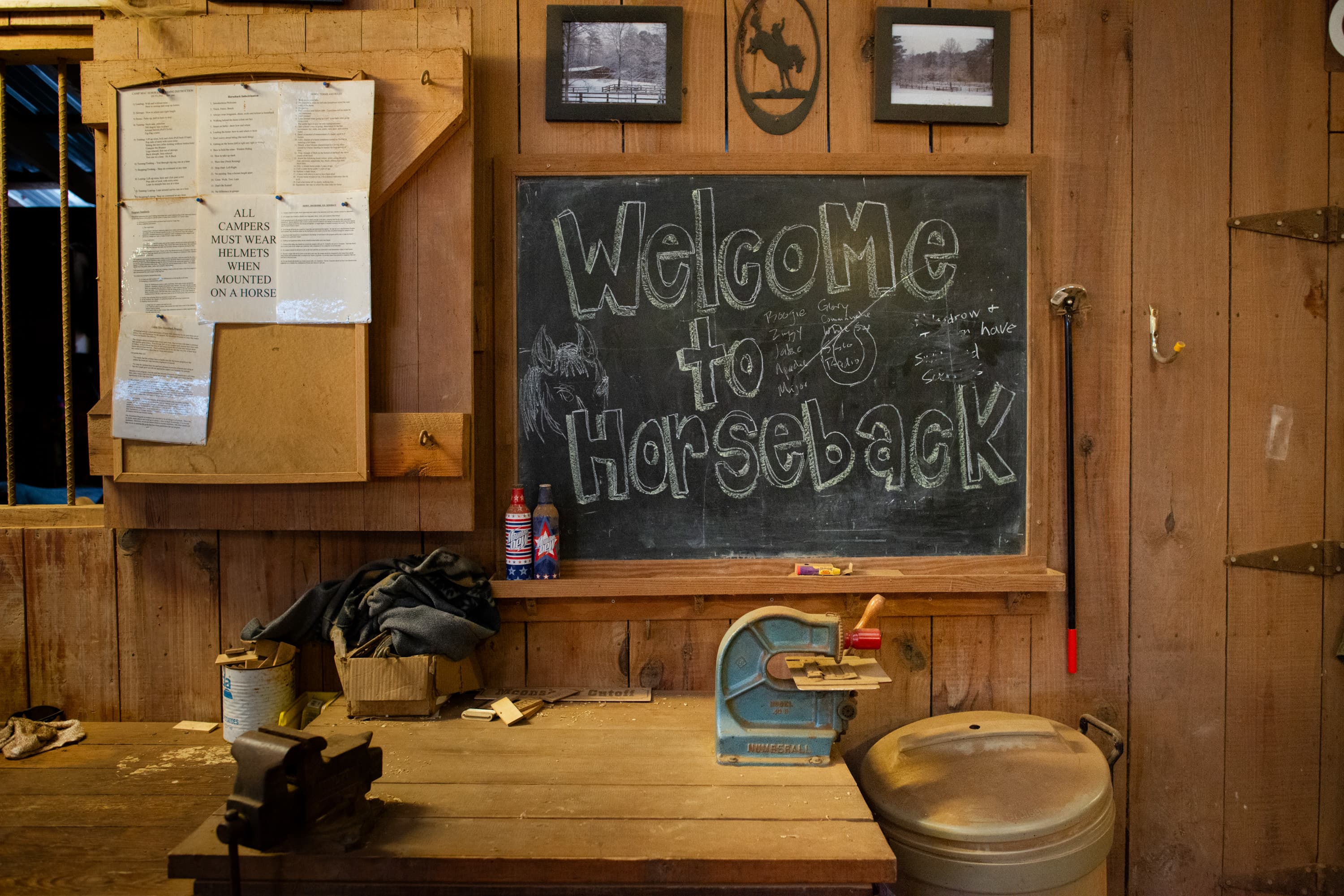 A wooden wall with a blackboard reading "Welcome to Horseback." Above the blackboard are two framed pictures and a horse emblem. To the left, a bulletin board has multiple documents and a sign that says "ALL CAMPERS MUST WEAR HELMETS WHEN MOUNTED ON A HORSE." Below, a workbench is cluttered with items like bottles, clothing, a small tool, and a vice.