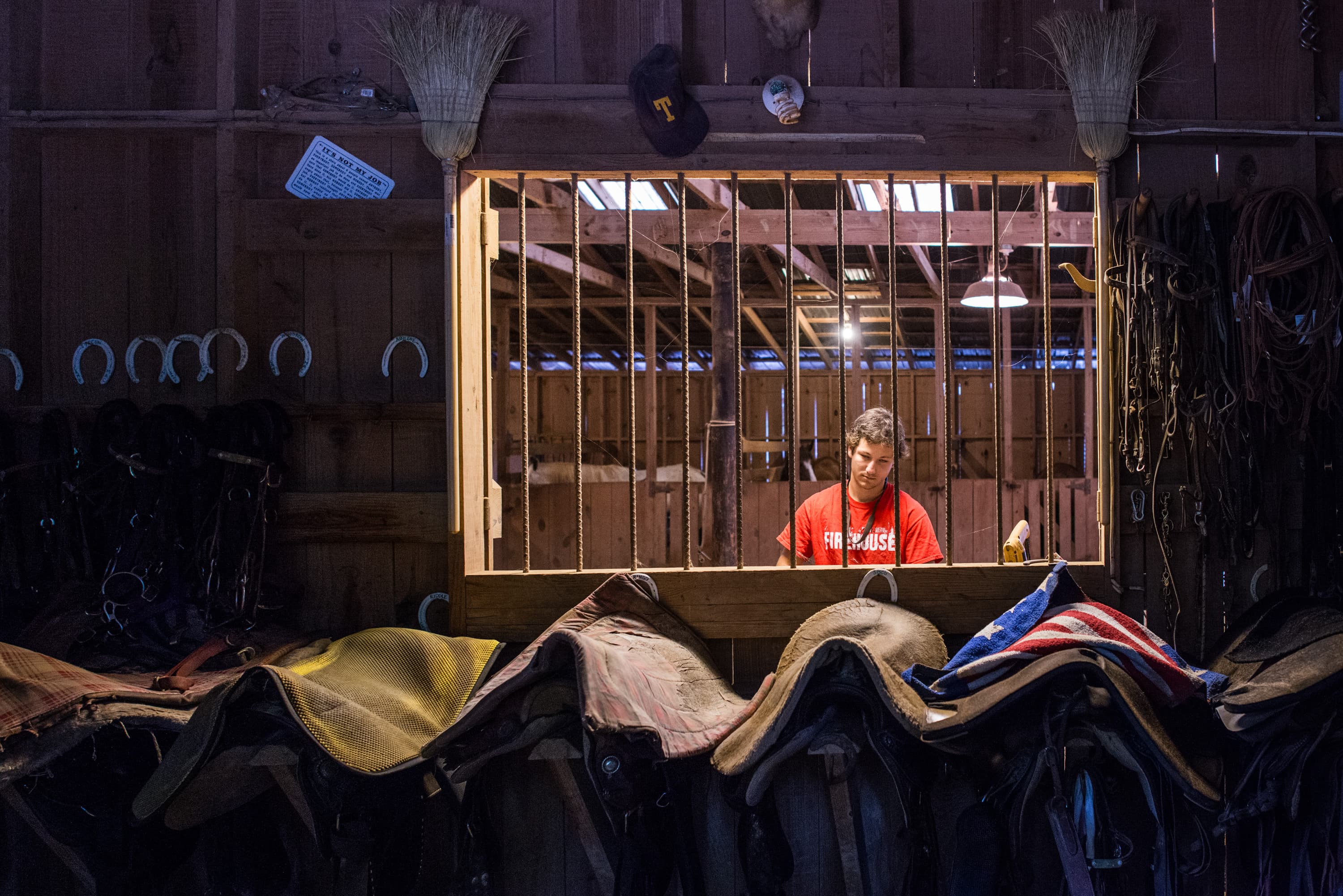 A person wearing a red "Firehouse" shirt is standing inside a wooden structure, seen through a window with bars. Below the window are saddles and horse equipment. Horseshoes are hanging on the wall, and various items like brooms and hats are displayed above.