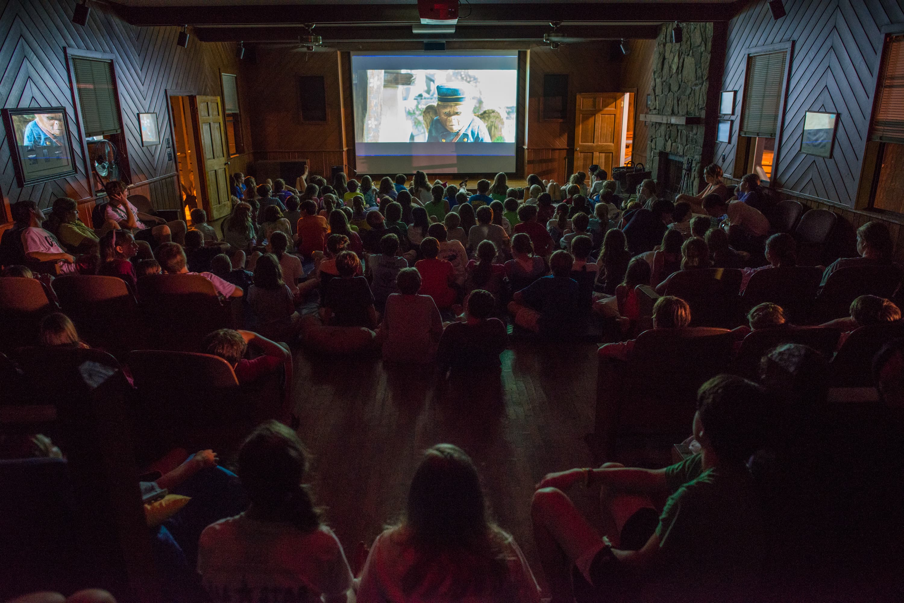 A group of people watching a movie on a screen in a dimly lit room with chairs and a wood-paneled interior.